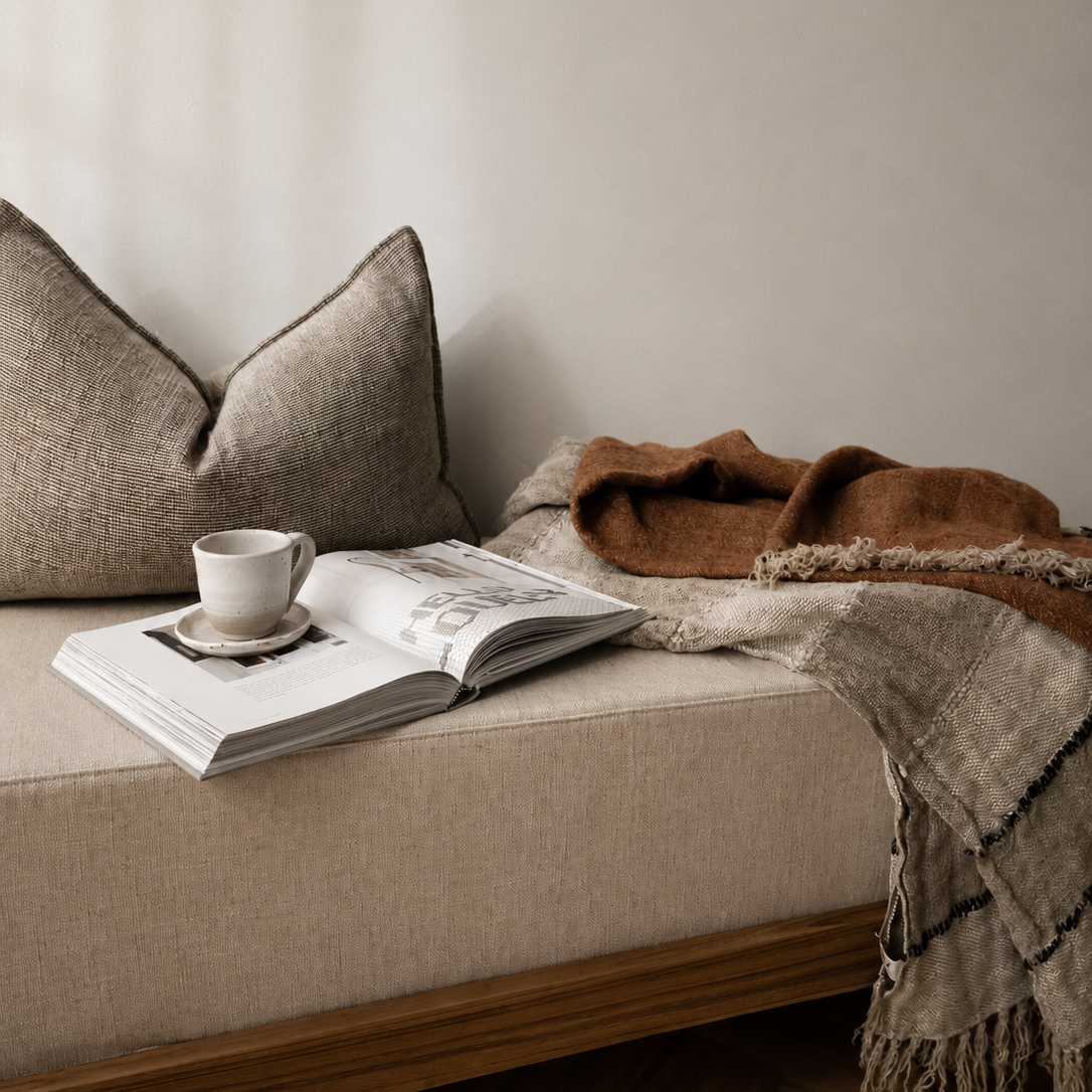 Beige sofa with a book, cup, and blanket on a wooden floor.